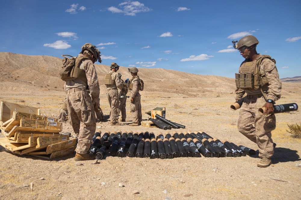 Military personnel in desert camouflage uniforms are handling and organizing what appear to be mortar rounds or artillery shells in an arid desert training environment. Multiple rounds are laid out in rows on the ground, with opened wooden ammunition crates visible to the left. The setting appears to be a military training range based on the terrain and organized nature of the activity.