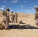 Military personnel in desert camouflage uniforms are handling and organizing what appear to be mortar rounds or artillery shells in an arid desert training environment. Multiple rounds are laid out in rows on the ground, with opened wooden ammunition crates visible to the left. The setting appears to be a military training range based on the terrain and organized nature of the activity.