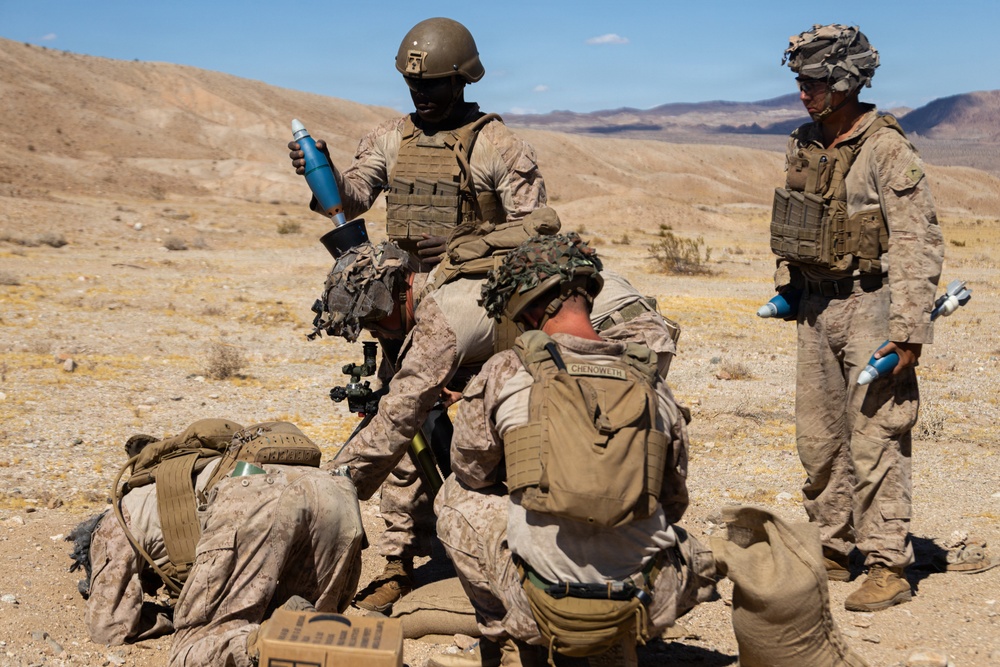 A group of military personnel in desert camouflage uniforms are gathered in an arid desert training environment. Several soldiers are crouched or kneeling around what appears to be a mortar or weapons system on the ground, while others stand nearby holding water bottles. The setting appears to be a desert training exercise based on the terrain and equipment visible.