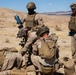 A group of military personnel in desert camouflage uniforms are gathered in an arid desert training environment. Several soldiers are crouched or kneeling around what appears to be a mortar or weapons system on the ground, while others stand nearby holding water bottles. The setting appears to be a desert training exercise based on the terrain and equipment visible.