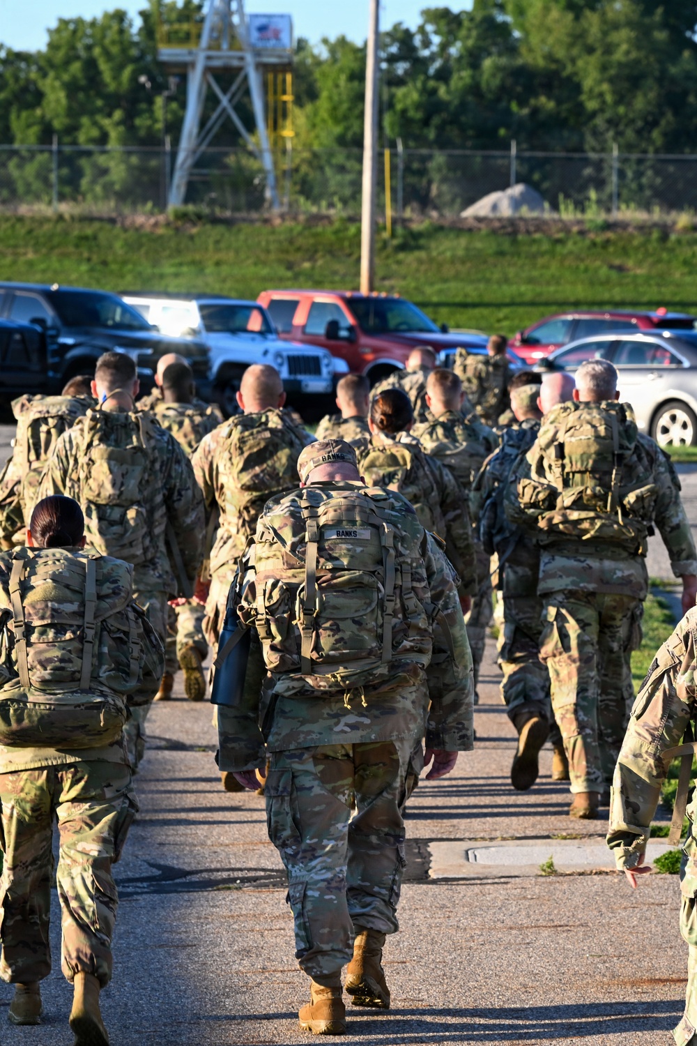 A large group of military personnel in camouflage uniforms and full rucksacks are marching away from the camera along a paved path. The soldiers appear to be conducting a ruck march or formation movement on what appears to be a military installation. A parking lot with civilian vehicles and a training tower structure are visible in the background.