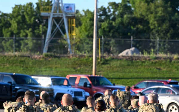 A large group of military personnel in camouflage uniforms and full rucksacks are marching away from the camera along a paved path. The soldiers appear to be conducting a ruck march or formation movement on what appears to be a military installation. A parking lot with civilian vehicles and a training tower structure are visible in the background.