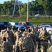 A large group of military personnel in camouflage uniforms and full rucksacks are marching away from the camera along a paved path. The soldiers appear to be conducting a ruck march or formation movement on what appears to be a military installation. A parking lot with civilian vehicles and a training tower structure are visible in the background.