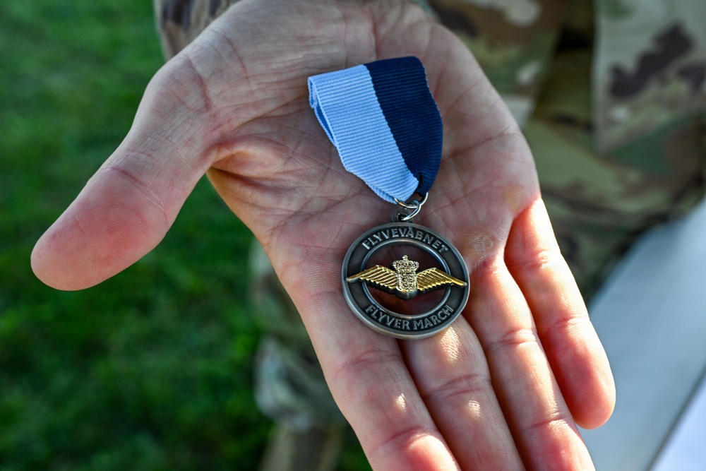 A hand, belonging to a person wearing military camouflage uniform, holds a circular medal with a blue and white ribbon. The medal bears the inscriptions 'FLYVEVÅBNET' (Royal Danish Air Force) and 'FLYVER MARCH,' featuring a gold winged emblem with a crown. This appears to be a Danish Air Force marching medal awarded for participation in a military march event.