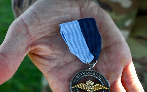 A hand, belonging to a person wearing military camouflage uniform, holds a circular medal with a blue and white ribbon. The medal bears the inscriptions 'FLYVEVÅBNET' (Royal Danish Air Force) and 'FLYVER MARCH,' featuring a gold winged emblem with a crown. This appears to be a Danish Air Force marching medal awarded for participation in a military march event.
