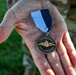 A hand, belonging to a person wearing military camouflage uniform, holds a circular medal with a blue and white ribbon. The medal bears the inscriptions 'FLYVEVÅBNET' (Royal Danish Air Force) and 'FLYVER MARCH,' featuring a gold winged emblem with a crown. This appears to be a Danish Air Force marching medal awarded for participation in a military march event.