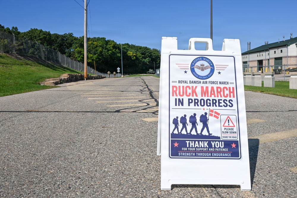 A white A-frame sandwich board sign is prominently displayed on a road announcing a 'Royal Danish Air Force March - Ruck March In Progress.' The sign features the Danish flag, silhouettes of marching soldiers with rucksacks, and a safety message asking drivers to slow down and share the road. The setting appears to be a military installation on a clear, sunny day.