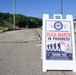 A white A-frame sandwich board sign is prominently displayed on a road announcing a 'Royal Danish Air Force March - Ruck March In Progress.' The sign features the Danish flag, silhouettes of marching soldiers with rucksacks, and a safety message asking drivers to slow down and share the road. The setting appears to be a military installation on a clear, sunny day.