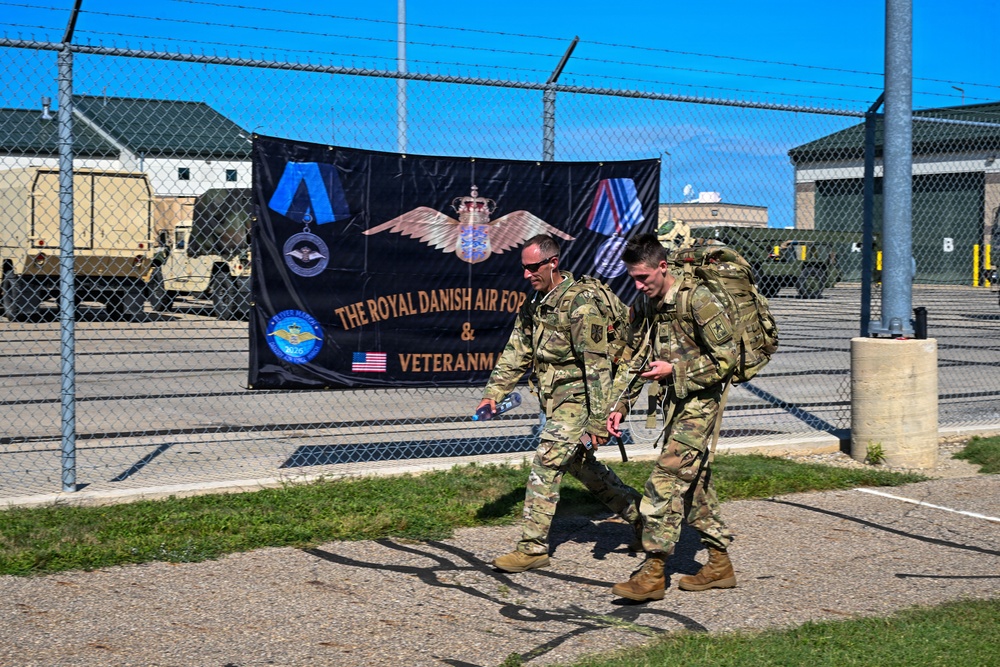 Two military personnel in camouflage uniforms and full tactical gear walk past a chain-link fence at what appears to be a military installation. A large black banner on the fence reads 'The Royal Danish Air Force &amp; Veteranm...' (partially obscured), featuring military insignia, medals, and an American flag. The background shows military vehicles, hangars, and open tarmac under a clear blue sky.