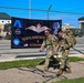 Two military personnel in camouflage uniforms and full tactical gear walk past a chain-link fence at what appears to be a military installation. A large black banner on the fence reads 'The Royal Danish Air Force &amp; Veteranm...' (partially obscured), featuring military insignia, medals, and an American flag. The background shows military vehicles, hangars, and open tarmac under a clear blue sky.