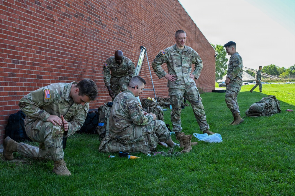 A group of U.S. Army soldiers in OCP (Operational Camouflage Pattern) uniforms are gathered on a grass lawn beside a red brick building. Several soldiers are crouching or sitting on the ground, appearing to organize or inspect gear and equipment. The scene suggests a pre- or post-activity staging area, likely on a military installation.