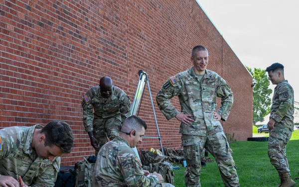 A group of U.S. Army soldiers in OCP (Operational Camouflage Pattern) uniforms are gathered on a grass lawn beside a red brick building. Several soldiers are crouching or sitting on the ground, appearing to organize or inspect gear and equipment. The scene suggests a pre- or post-activity staging area, likely on a military installation.