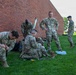 A group of U.S. Army soldiers in OCP (Operational Camouflage Pattern) uniforms are gathered on a grass lawn beside a red brick building. Several soldiers are crouching or sitting on the ground, appearing to organize or inspect gear and equipment. The scene suggests a pre- or post-activity staging area, likely on a military installation.