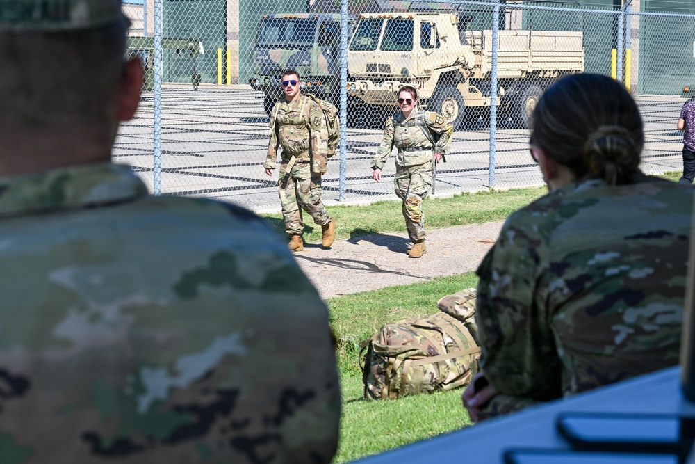 The image shows multiple military personnel in OCP (Operational Camouflage Pattern) uniforms at what appears to be a military installation or motor pool area. Two soldiers wearing sunglasses are walking toward the camera along a pathway, while two others are visible in the foreground with their backs turned. Military vehicles, including what appears to be a tactical truck, are visible behind a chain-link fence in the background.