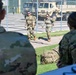 The image shows multiple military personnel in OCP (Operational Camouflage Pattern) uniforms at what appears to be a military installation or motor pool area. Two soldiers wearing sunglasses are walking toward the camera along a pathway, while two others are visible in the foreground with their backs turned. Military vehicles, including what appears to be a tactical truck, are visible behind a chain-link fence in the background.