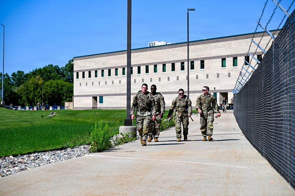 Four military personnel in camouflage uniforms are walking together along a concrete pathway adjacent to a chain-link fence topped with razor wire. They are on what appears to be a military installation or secure facility, with a large institutional building visible in the background. The scene suggests a routine patrol or movement between locations on the base.