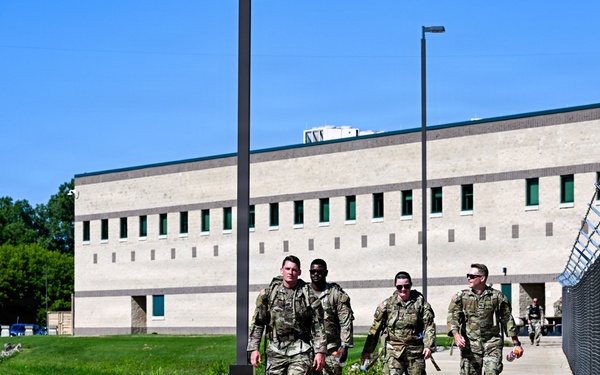 Four military personnel in camouflage uniforms are walking together along a concrete pathway adjacent to a chain-link fence topped with razor wire. They are on what appears to be a military installation or secure facility, with a large institutional building visible in the background. The scene suggests a routine patrol or movement between locations on the base.