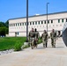 Four military personnel in camouflage uniforms are walking together along a concrete pathway adjacent to a chain-link fence topped with razor wire. They are on what appears to be a military installation or secure facility, with a large institutional building visible in the background. The scene suggests a routine patrol or movement between locations on the base.