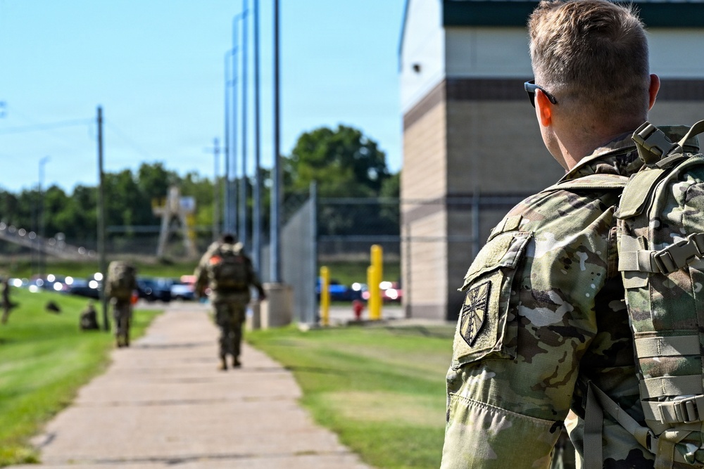 Military personnel in Operational Camouflage Pattern (OCP) uniforms are walking along a paved pathway near a building and fenced area. The foreground soldier is wearing a tactical backpack and sunglasses, with a unit patch visible on the left sleeve. Additional soldiers are visible in the background moving in the same direction.