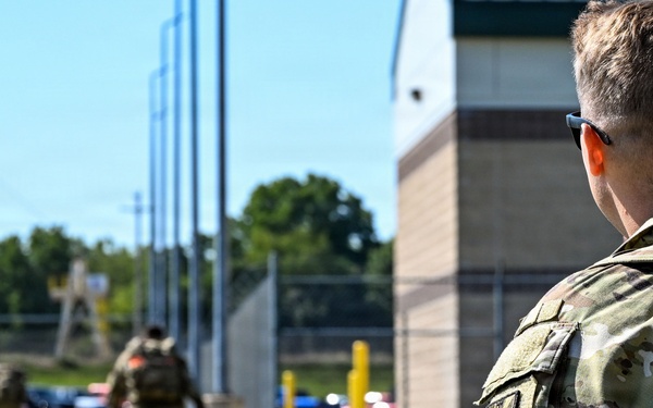 Military personnel in Operational Camouflage Pattern (OCP) uniforms are walking along a paved pathway near a building and fenced area. The foreground soldier is wearing a tactical backpack and sunglasses, with a unit patch visible on the left sleeve. Additional soldiers are visible in the background moving in the same direction.
