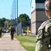Military personnel in Operational Camouflage Pattern (OCP) uniforms are walking along a paved pathway near a building and fenced area. The foreground soldier is wearing a tactical backpack and sunglasses, with a unit patch visible on the left sleeve. Additional soldiers are visible in the background moving in the same direction.