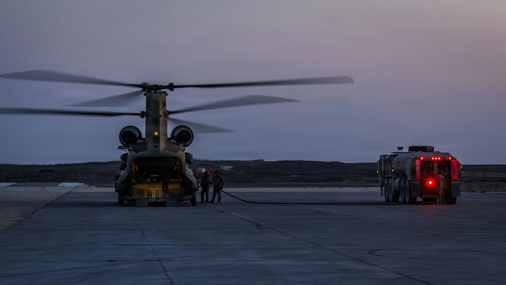 A military CH-47 Chinook tandem-rotor helicopter is being refueled on a tarmac at dusk or dawn. A fuel tanker truck is connected to the aircraft via a hose, with several personnel visible near the rear ramp of the helicopter. The low-light conditions and muted sky suggest early morning or evening operations.