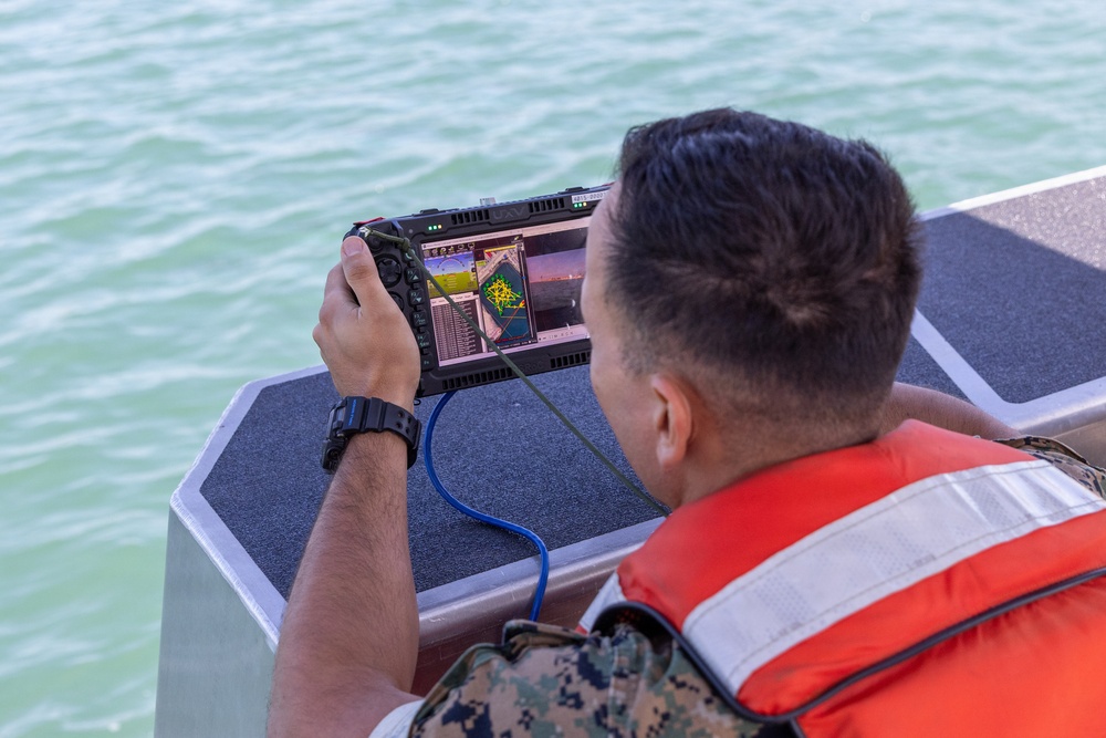 A military personnel member wearing camouflage uniform and an orange life vest is operating a ruggedized handheld drone or UAV ground control station while aboard a watercraft. The device displays what appears to be navigation/mapping data and possibly a live camera feed. The setting is on open water, suggesting a maritime reconnaissance or surveillance operation.