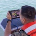 A military personnel member wearing camouflage uniform and an orange life vest is operating a ruggedized handheld drone or UAV ground control station while aboard a watercraft. The device displays what appears to be navigation/mapping data and possibly a live camera feed. The setting is on open water, suggesting a maritime reconnaissance or surveillance operation.