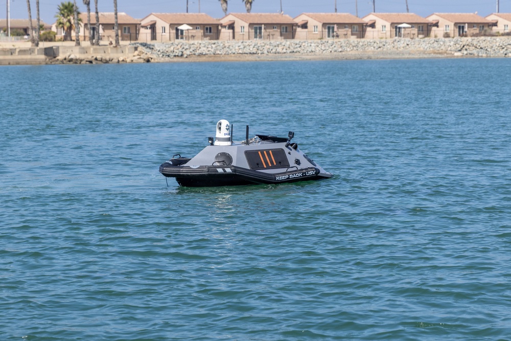 The image shows an unmanned surface vehicle (USV) operating on open water near a coastal residential area. The vessel is gray and black with orange markings and bears the text 'KEEP BACK - USV', indicating it is an autonomous or remotely operated watercraft. Palm trees and uniform tan-colored buildings are visible in the background, suggesting a Southern California or similar coastal environment.