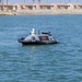The image shows an unmanned surface vehicle (USV) operating on open water near a coastal residential area. The vessel is gray and black with orange markings and bears the text 'KEEP BACK - USV', indicating it is an autonomous or remotely operated watercraft. Palm trees and uniform tan-colored buildings are visible in the background, suggesting a Southern California or similar coastal environment.