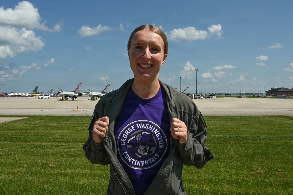 A smiling woman stands on an airfield tarmac area, proudly displaying a purple 'George Washington Continentals' t-shirt beneath an olive green flight jacket. In the background, several U.S. Air Force Thunderbirds aircraft are visible parked on the flight line, suggesting this was taken at an air show. The setting appears to be a sunny summer day at a military airfield.