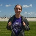 A smiling woman stands on an airfield tarmac area, proudly displaying a purple 'George Washington Continentals' t-shirt beneath an olive green flight jacket. In the background, several U.S. Air Force Thunderbirds aircraft are visible parked on the flight line, suggesting this was taken at an air show. The setting appears to be a sunny summer day at a military airfield.