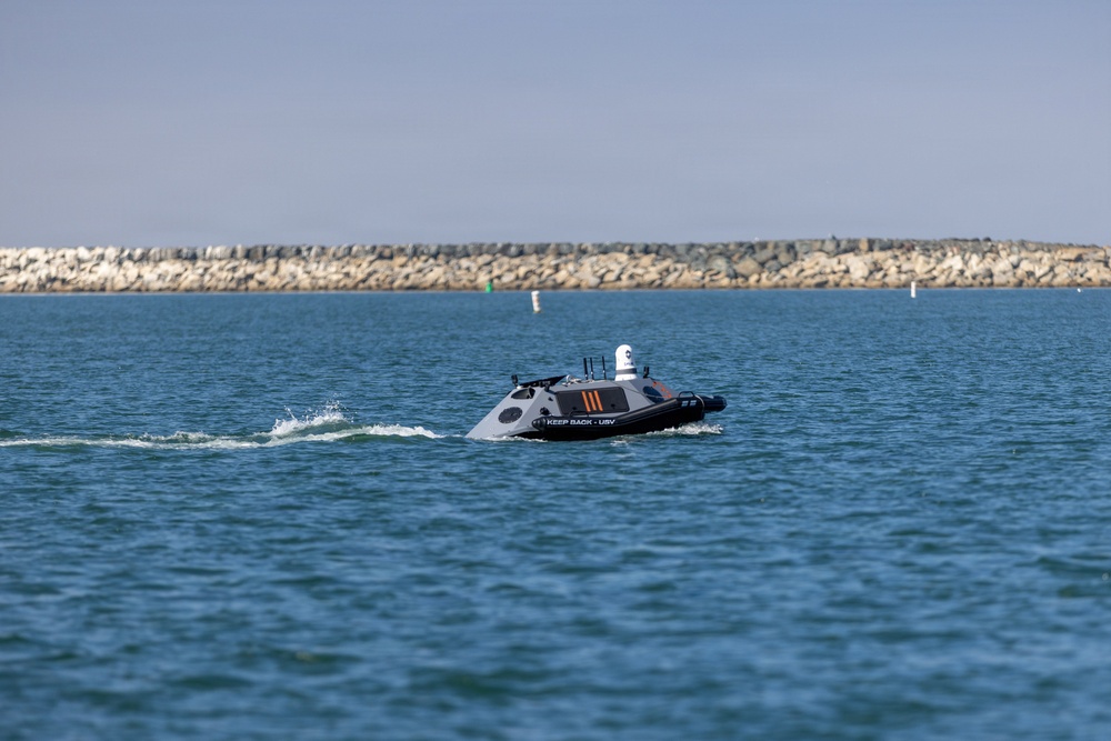 The image shows an unmanned surface vehicle (USV) navigating open water near a rocky breakwater. The dark-hulled autonomous vessel is equipped with sensors and a turret-mounted camera system on top, and bears the text 'KEEP BACK - USV' on its hull. The scene appears to be a coastal or harbor environment under clear skies.