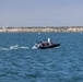 The image shows an unmanned surface vehicle (USV) navigating open water near a rocky breakwater. The dark-hulled autonomous vessel is equipped with sensors and a turret-mounted camera system on top, and bears the text 'KEEP BACK - USV' on its hull. The scene appears to be a coastal or harbor environment under clear skies.