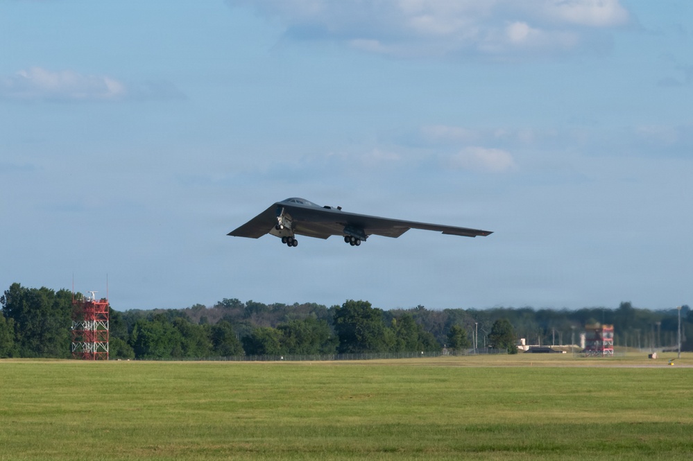 A B-2 Spirit stealth bomber is captured during takeoff or low-altitude flight over a military airfield. The aircraft's distinctive flying-wing design is clearly visible with landing gear still deployed. The setting appears to be a well-maintained military air base with green grass, tree lines, and ground infrastructure visible in the background.