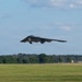 A B-2 Spirit stealth bomber is captured during takeoff or low-altitude flight over a military airfield. The aircraft's distinctive flying-wing design is clearly visible with landing gear still deployed. The setting appears to be a well-maintained military air base with green grass, tree lines, and ground infrastructure visible in the background.