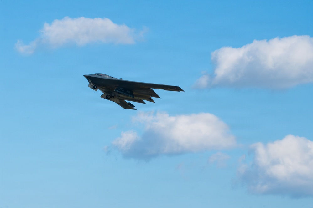 A B-2 Spirit stealth bomber is captured in flight against a blue sky with scattered white clouds. The aircraft is photographed from a slightly below-and-behind angle, clearly showing its distinctive flying wing design and dark gray coloration. Ordnance or external stores appear to be visible beneath the fuselage.