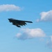 A B-2 Spirit stealth bomber is captured in flight against a blue sky with scattered white clouds. The aircraft is photographed from a slightly below-and-behind angle, clearly showing its distinctive flying wing design and dark gray coloration. Ordnance or external stores appear to be visible beneath the fuselage.