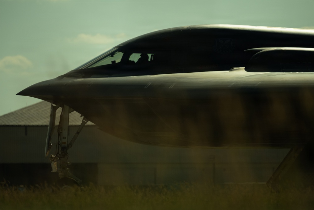A close-up, low-angle photograph of what appears to be a B-2 Spirit stealth bomber on the ground, captured in dramatic lighting with a moody, dark aesthetic. The aircraft's distinctive flying-wing design, smooth blended fuselage, and cockpit canopy are clearly visible. A building consistent with a military hangar is partially visible in the background.
