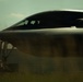 A close-up, low-angle photograph of what appears to be a B-2 Spirit stealth bomber on the ground, captured in dramatic lighting with a moody, dark aesthetic. The aircraft's distinctive flying-wing design, smooth blended fuselage, and cockpit canopy are clearly visible. A building consistent with a military hangar is partially visible in the background.