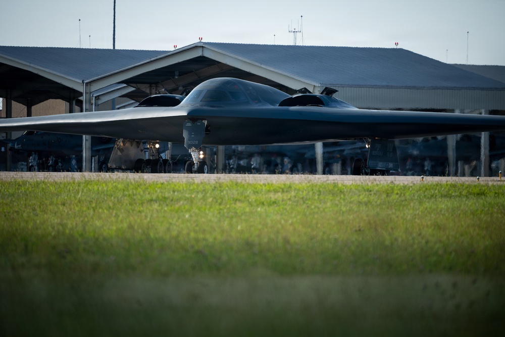 A B-2 Spirit stealth bomber is taxiing on a runway apron in front of a large military hangar facility. The aircraft's distinctive flying-wing design and dark radar-absorbing coating are clearly visible. Additional aircraft and ground personnel are visible in the background under the hangar structure.
