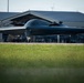A B-2 Spirit stealth bomber is taxiing on a runway apron in front of a large military hangar facility. The aircraft's distinctive flying-wing design and dark radar-absorbing coating are clearly visible. Additional aircraft and ground personnel are visible in the background under the hangar structure.