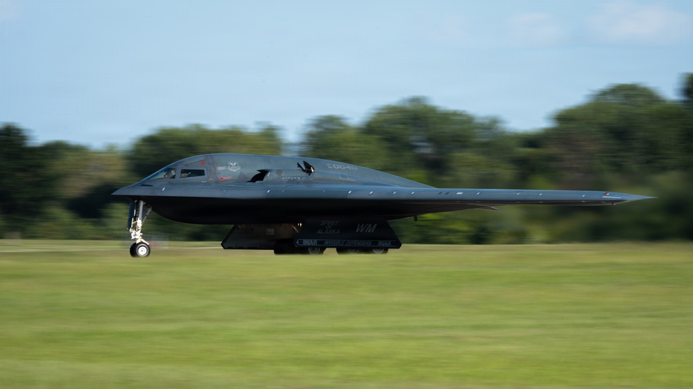 A B-2 Spirit stealth bomber is captured in motion on a runway, likely during takeoff or landing. The aircraft is painted matte black and displays visible markings including 'WM' and 'ALASKA' on its underside. Motion blur on the background trees and grass emphasizes the aircraft's speed.