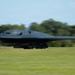 A B-2 Spirit stealth bomber is captured in motion on a runway, likely during takeoff or landing. The aircraft is painted matte black and displays visible markings including 'WM' and 'ALASKA' on its underside. Motion blur on the background trees and grass emphasizes the aircraft's speed.