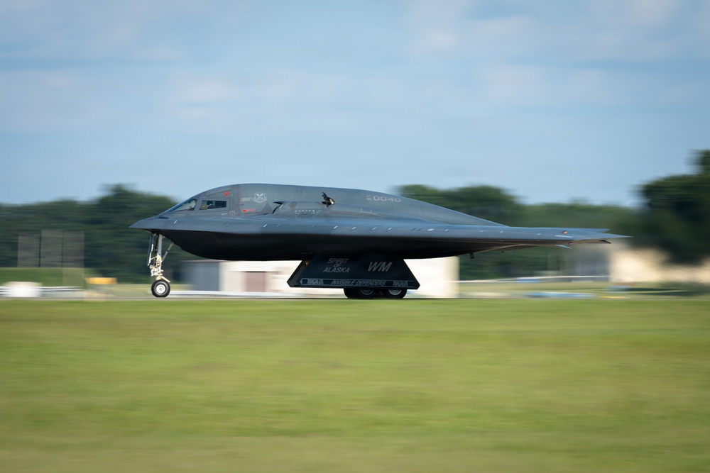 A B-2 Spirit stealth bomber is captured during a ground roll on a runway, appearing to be in the process of taking off or landing. The aircraft is painted matte black and bears visible markings including 'Spirit of Alaska' and the tail code 'WM' along with the serial number '20040'. Motion blur on the background indicates the aircraft is moving at speed.