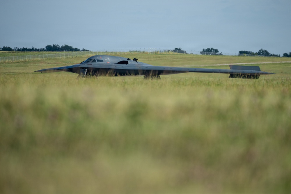 A B-2 Spirit stealth bomber is photographed on the ground at an airfield, viewed through blurred foreground grass that creates a low-angle perspective. The aircraft's distinctive flying-wing design and dark gray finish are clearly visible. The setting appears to be a military airfield with green grass, trees, and fencing visible in the background.