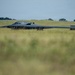 A B-2 Spirit stealth bomber is photographed on the ground at an airfield, viewed through blurred foreground grass that creates a low-angle perspective. The aircraft's distinctive flying-wing design and dark gray finish are clearly visible. The setting appears to be a military airfield with green grass, trees, and fencing visible in the background.