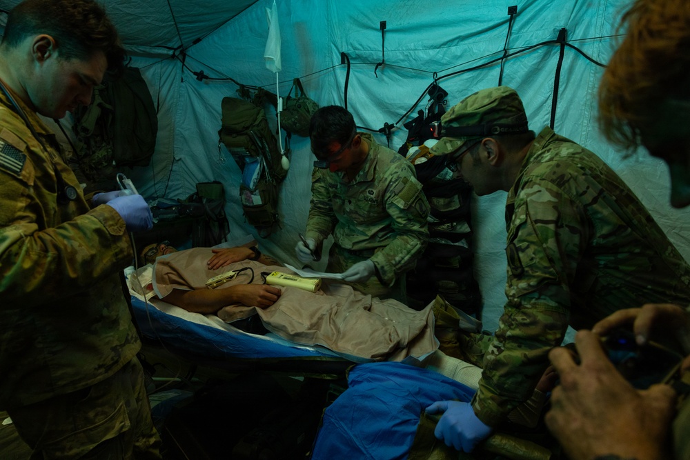 Military medical personnel in camouflage uniforms are treating a patient on a field stretcher inside a tactical tent structure. The scene appears to be a combat casualty care exercise or real field medical operation, with multiple personnel wearing gloves and using medical equipment. The low-light environment with teal/blue ambient lighting suggests a nighttime or simulated operational scenario.