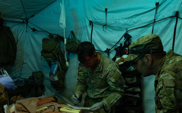 Military medical personnel in camouflage uniforms are treating a patient on a field stretcher inside a tactical tent structure. The scene appears to be a combat casualty care exercise or real field medical operation, with multiple personnel wearing gloves and using medical equipment. The low-light environment with teal/blue ambient lighting suggests a nighttime or simulated operational scenario.