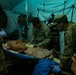 Military medical personnel in camouflage uniforms are treating a patient on a field stretcher inside a tactical tent structure. The scene appears to be a combat casualty care exercise or real field medical operation, with multiple personnel wearing gloves and using medical equipment. The low-light environment with teal/blue ambient lighting suggests a nighttime or simulated operational scenario.